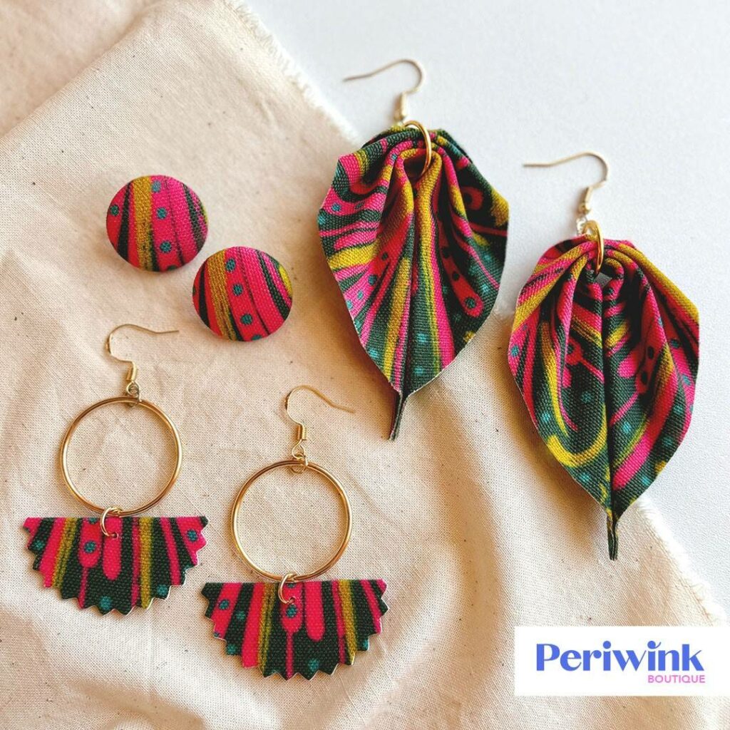 Three pairs of multicolor earrings rest of a cream colored cloth and white surface. One pair are button earrings, another pair are dangling leaf-shaped, and the third pair are hoops with fabric dangling from the bottom center. 