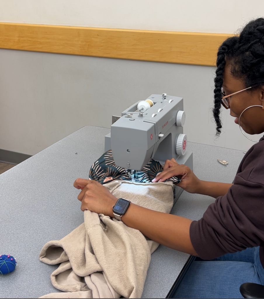 Arlette sews the satin to the neckline of her hoodie using a grey sewing machine. Her hands are placed on both ends of the hoodie to keep the fabric flat.