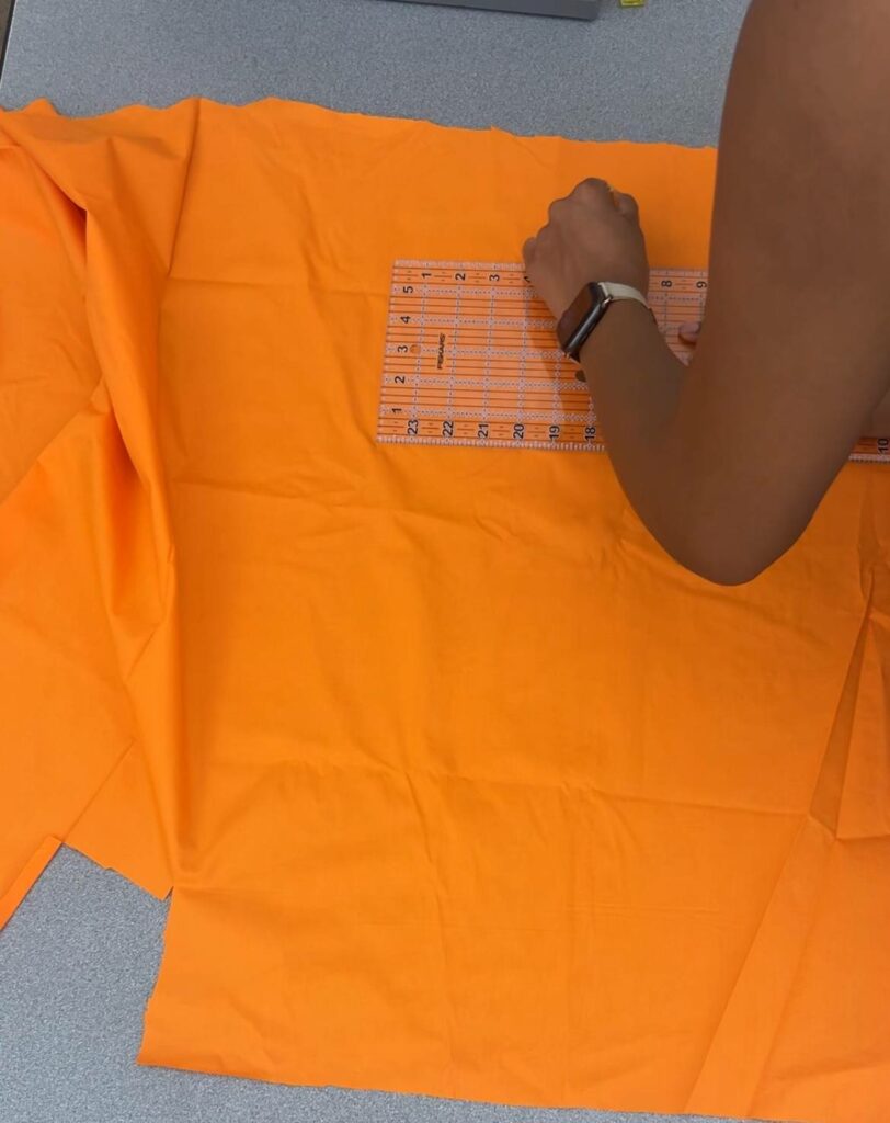 Using her sewing ruler, Arlette traces out her measurements onto her orange scrap fabric.