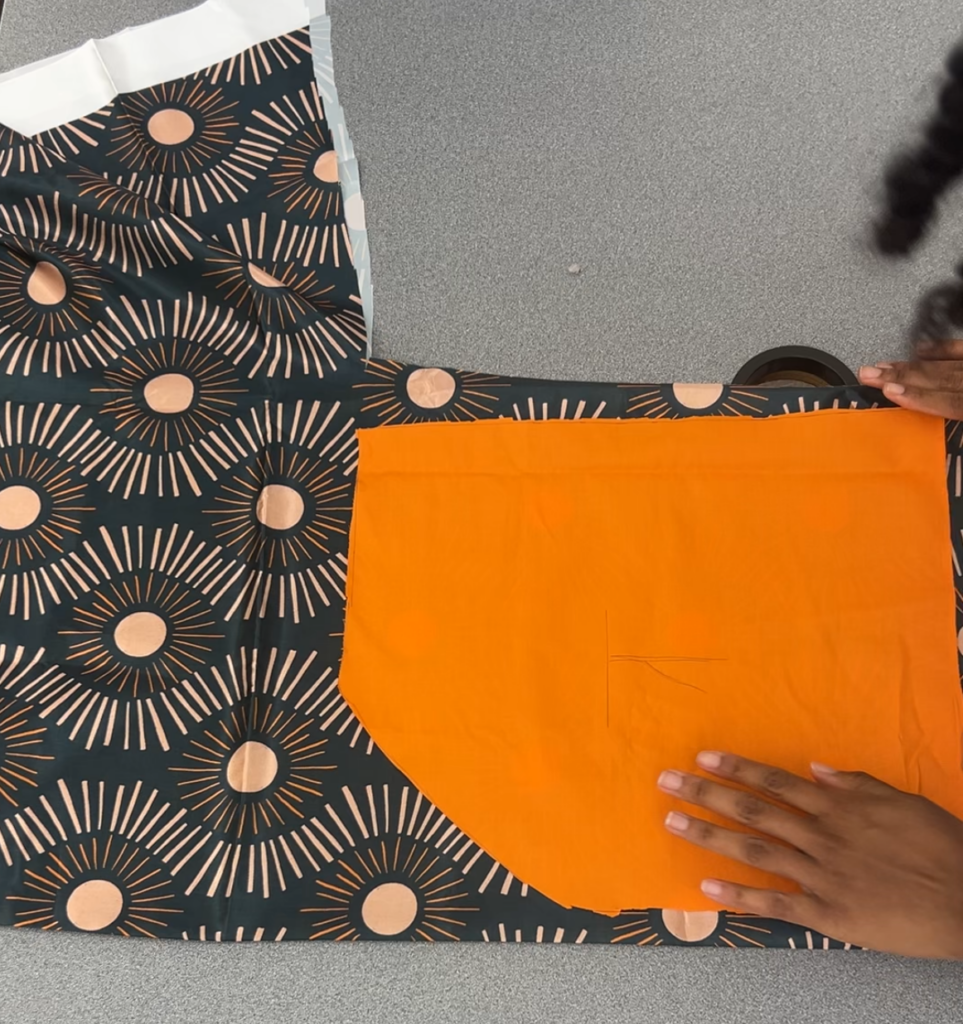 Arlette lays the orange template on top of her satin fabric. The fabric is dark blue with tan lines going in a wavy pattern. Darker brown lines surround circles like a sun and its rays. The fabric lays on a grey table.