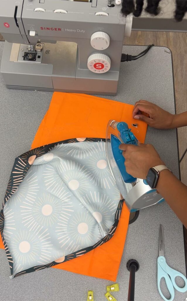 Arlette is ironing the seams of the satin hood using a blue iron. The orange fabric acts as an ironing cloth for the satin. Her other supplies lay scattered about the table.