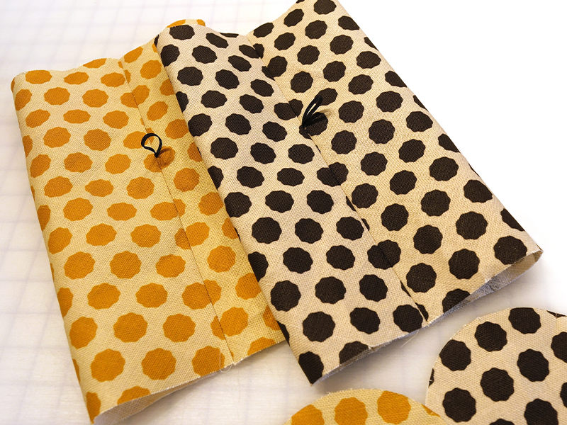 The fabric bodies of both the inside and outside bag and round bag bottoms lay on a white cutting mat with a black grid. Some of the fabric has a white background with black dots and some of the fabric has gold dots on a light yellow background.