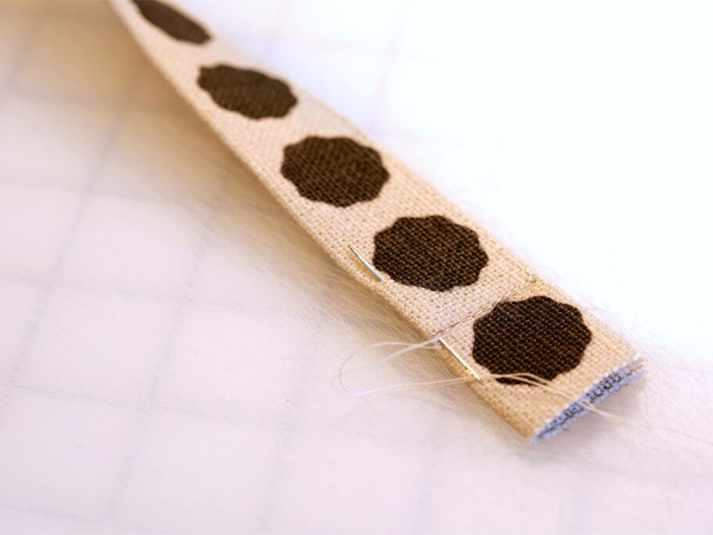 A needle has been inserted vertically on the left handside of a long thing piece of fabric that is white with black dots. The needle is being used to sew in fabric ends.