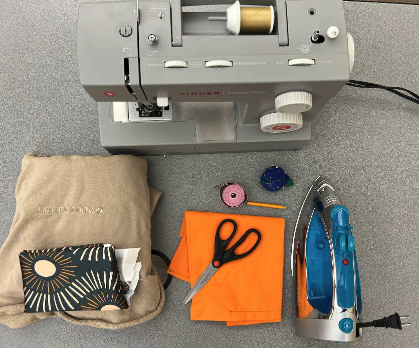 Materials to make a satin lined hoodie lay on a grey table including a sewing machine, measuring tape, scissors, fabric, iron, pencil, pin cushion and sewing pins, satin fabric, and hoodie.