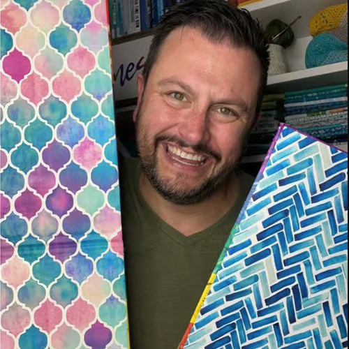 Mathew Boudreaux holds up backdrops made out of cardboard and smiles. The design on the backdrop to the left has a watercolor tile design. The design on the backdrop to the right is a blue and white herringbone.