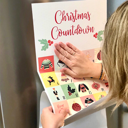 A person is sticking a Peel and Stick advent calendar that says "Christmas Countdown" at the top to the front of their refrigerator.