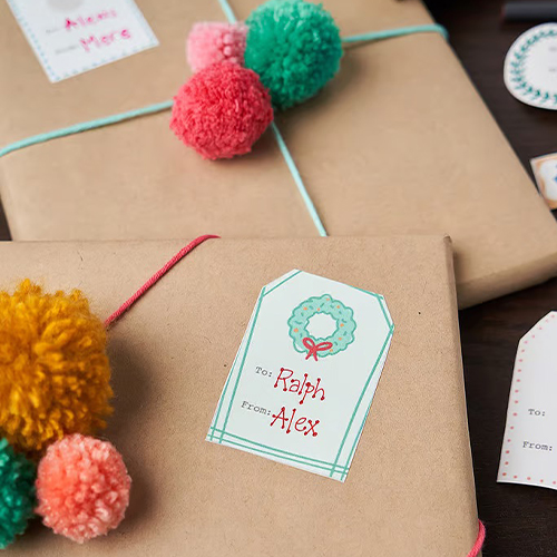 Small Peel and Stick gift tags adorn packages wrapped in brown paper and tied with yarn and jewel-toned pom poms.