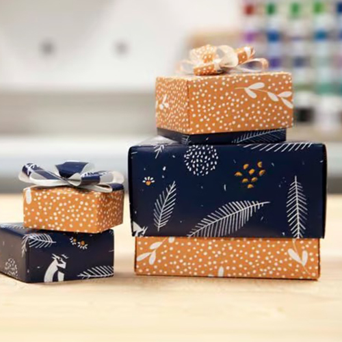 Two small stacks of boxes made out of Prepasted Removable wallpaper lay on a wooden table. Some boxes are a dark yellow with white dots and small white twigs. Other boxes have a dark blue background with white leaves.