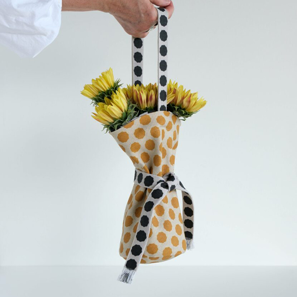 A hand holds a gift bag filled with yellow flowers. The bag has two layers, an outer layer in fabric that has gold dots on a yellow background and an inner layer with black dots on a white background. The bag’s tie in the middle and wrist strap are also black dots with a white background.