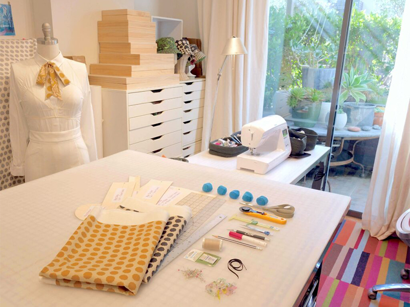 A sewing room with tutorial materials laid out on a cutting table. A sewing machine is nearby and a dress form wearing a white shirt with a yellow-and cream skinny tie is to the left. Outside the window to the right is a patio filled with small rocks and succulents in pots.