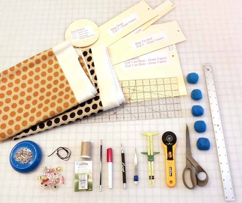 Materials for the wine bag tutorial are laid out on a white cutting table with a black grid. A rotary cutter, several templates for the pattern pieces, blue and green pattern weights, pins, scissors, a ruler, and two folded pieces of fabric (one that has gold dots on light yellow and one that has black dots on white) are on the table too along with a few other materials for the tutorial that are in the list below.
