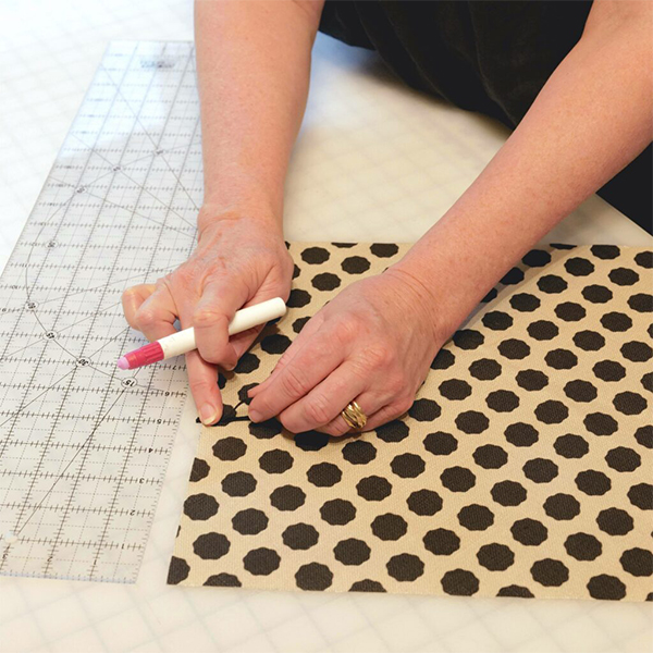 Cinne is basting a thin black elastic loop on to the top of fabric that has black dots and a white background. A large clear ruler is the left and the fabric is on a white cutting board with a black grid.