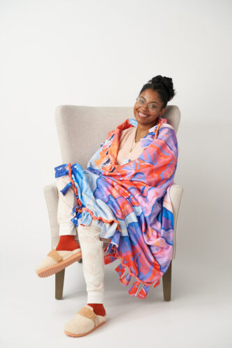 Kristina sitting in a grey chair, wrapped in a fleece blanket in front of a wihte background.