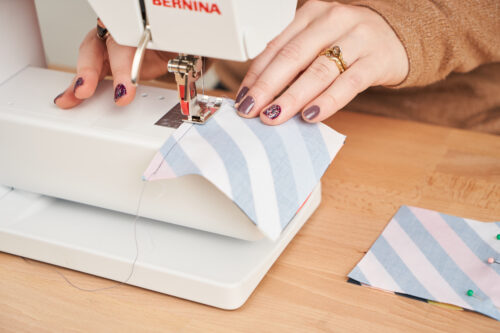 Hands sewing striped fabric design side down through a sewing machine on a wooden surface.