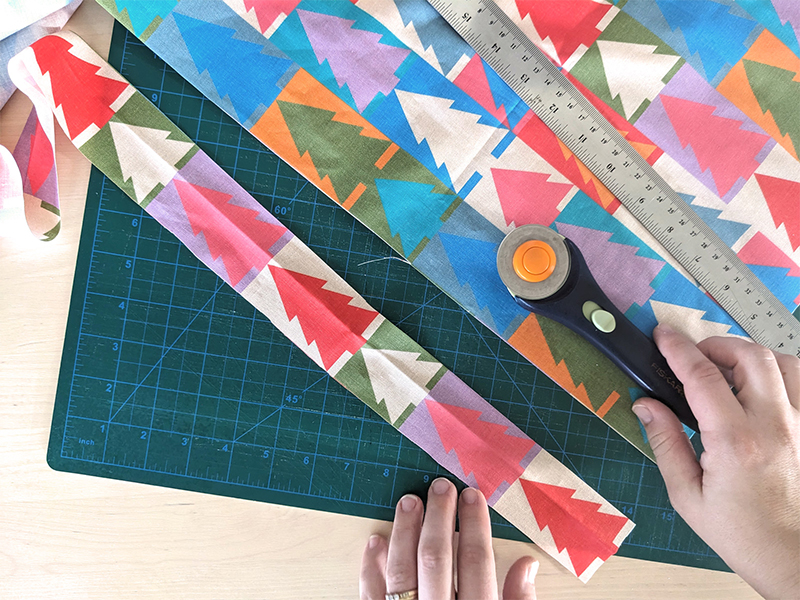 Lisa has cut one strip of fabric lengthwise in a long 2” strip. The fabric features a design with white and jewel tone repeating Christmas trees. The fabric is on a green cutting mat and Lisa is holding a rotary cutter at the photo’s bottom right.