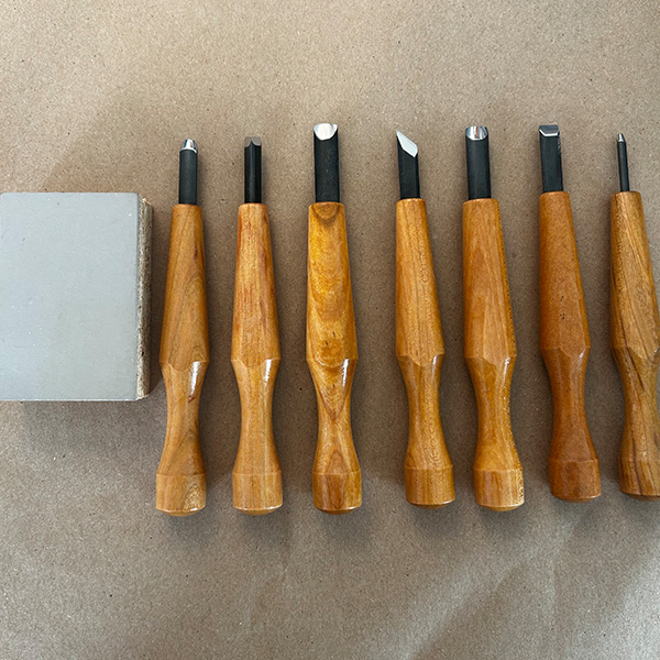 Seven wooden linoleum cutters lay beside a linoleum rectangle on a brown surface.