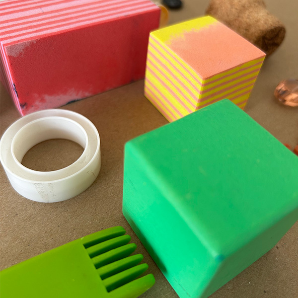 Found items like a sponge, tape cylinder, green block, and wine cork are on a brown surface.
