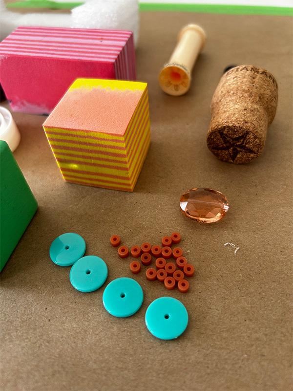Beads, a jewel, wine cork, and sponges rest on a brown surface.