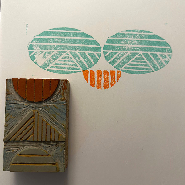 A teal and orange block print design is beside the linoleum block, on a white sheet of paper.