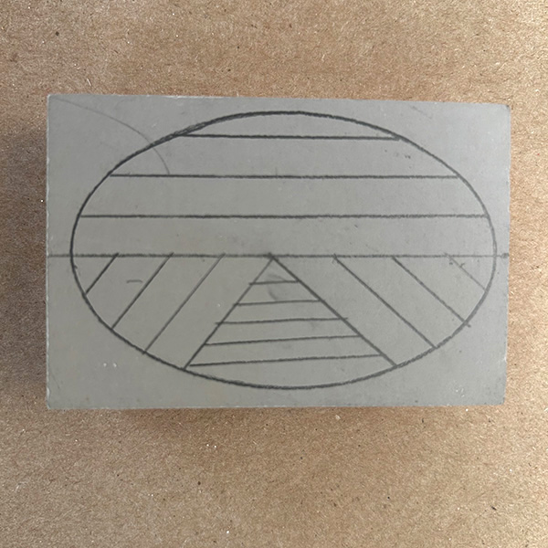 A grey rectangle of linoleum rests on a brown surface. A circle with lines going in different directions is drawn on the linoleum.