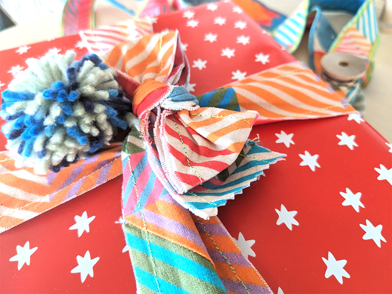 Close up of the top of a present wrapped in red wrapping paper with small white stars. Ribbon wrapped around the present has been made out of a 2”-wide fabric strips sewn together. The ribbon features a design with small squares of coordinating colors in varying pairings. A blue-and-gray pom pom has been tied to the ribbon’s bow.