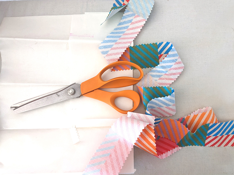 A long strip of 2”-wide cotton fabric featuring a design with small squares of coordinating colors in varying jewel tone pairings has been cut out with pinking shears. It is laying on a white surface. A pink or orange-handled pinking shears is laying on top of the fabric strip.