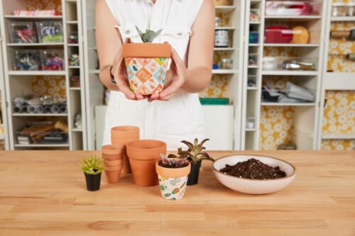 Alexa holding a plant pot covered with a fabric design. On a wooden table are the other supplies and plant pots waiting to be covered.