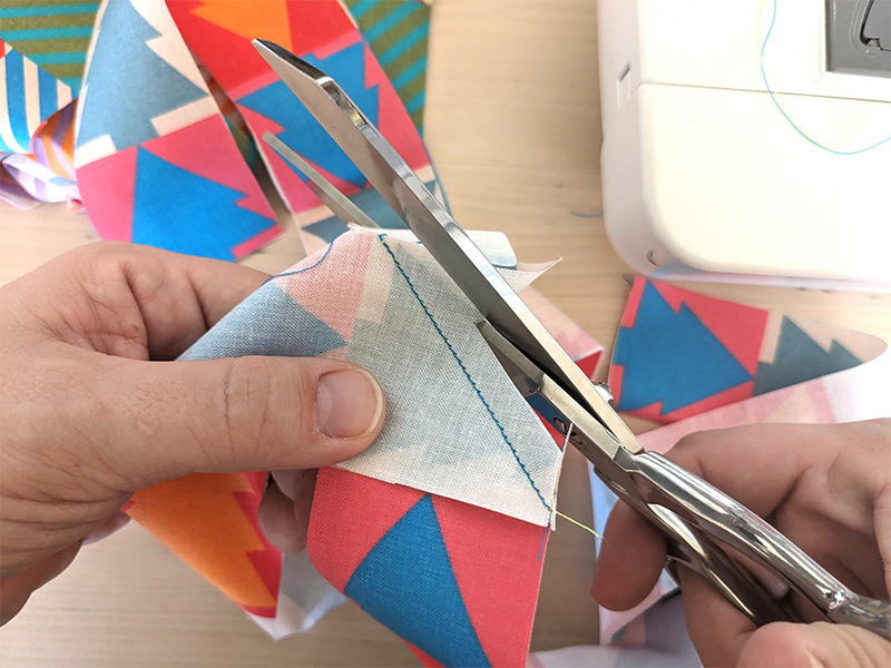 Lisa is cutting extra fabric left over from when two top corners of perpendicular strips of fabric were sewn together at a 45-degree angle. She cutting off the small extra triangles of fabric that are no longer needed.