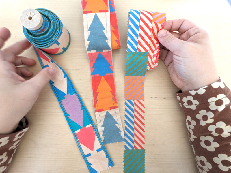 Examples of three different fabric ribbon. The two left-hand ribbons are made with a design featuring jewel-tone and white Christmas trees. The ribbon on the right has a design with small squares of coordinating colors in varying pairings, also in jewel-tone designs. The ribbon on the right has serged edges, the middle ribbon has sewn together edges and the ribbon on the right has pinking shear edges.