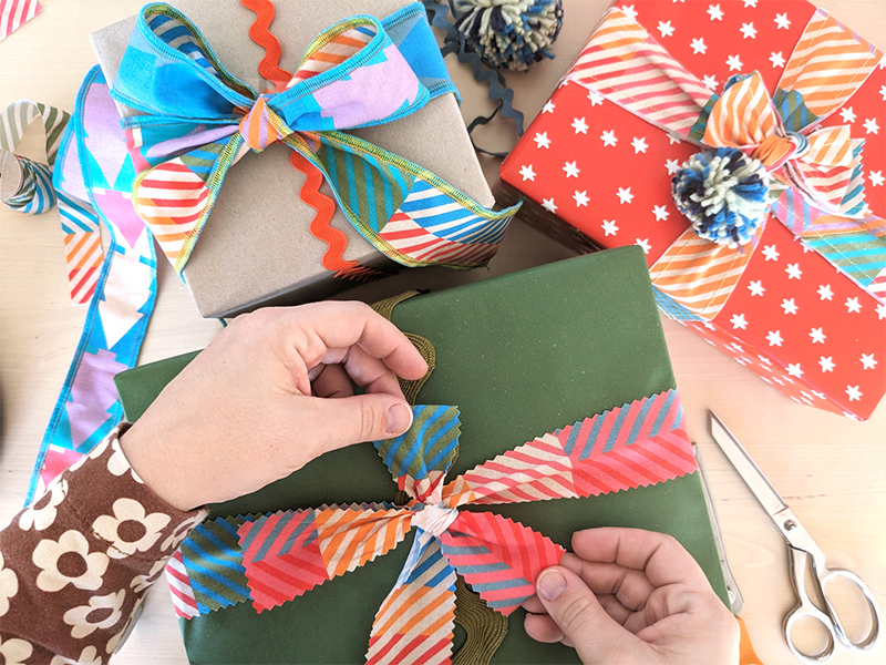 Three presents are on a table, all wrapped with fabric ribbons tied in bows. Two hands are tying the bow on top the present toward the bottom middle of the image. The bottom middle and right present are tied with fabric ribbon with a design featuring small squares of coordinating colors in varying pairings. The present to the left has been wrapped with ribbon with the small squares design on one side and a Christmas tree repeating design on the other.