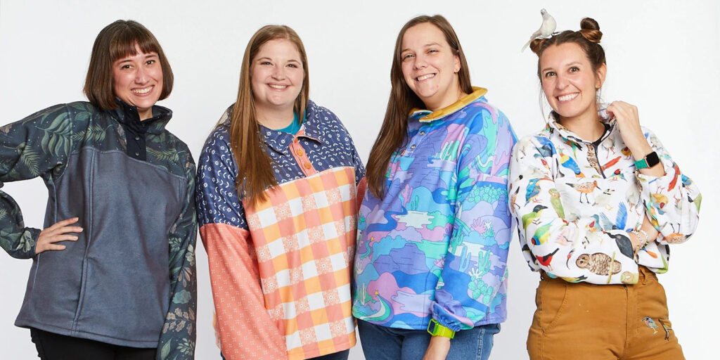 Four women are wearing fleece pullovers in different Spoonflower designs and standing in front of a white background. They are smiling and looking at the camera.