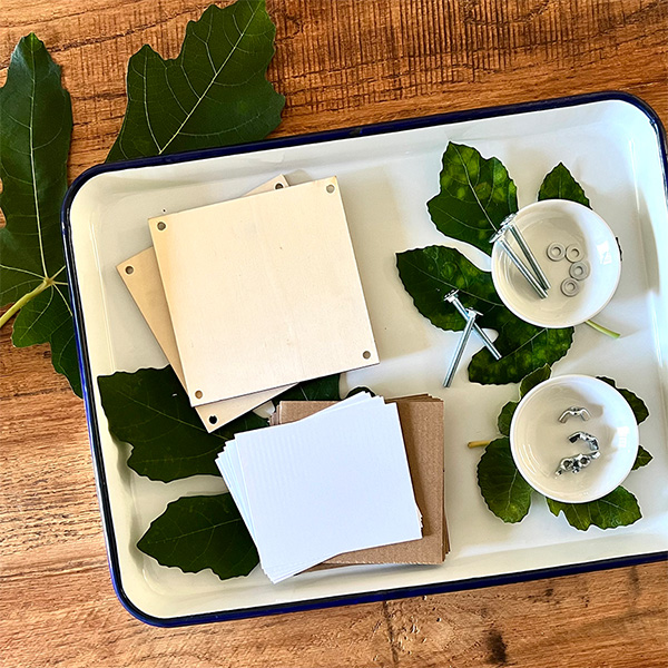 Two pieces of plywood, several pieces of cardboard and copy paper, four nuts, washers, and bolts sit on a white tray with green leaves. The tray is on a wooden surface with more leaves behind it.
