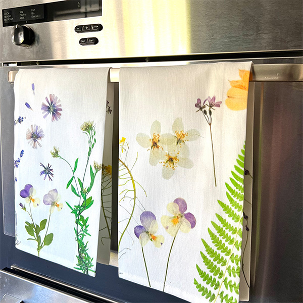 Two tea towels with various botanicals hang from the handle of a stainless steel appliance.