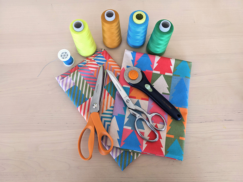 Materials for this project have been laid out on a wooden table: thread spools in yellow, orange, blue and green; two folded pieces of cotton fabric featuring designs in jewel tone colors, in both a Christmas tree repeating design and small squares of coordinating colors in varying pairings; scissors; pinking shears and a rotary cutter.