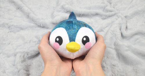 A penguin plushie rests in the palms of someones hands in front of a grey background.