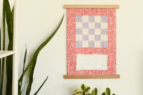 A quilted calendar hangs on a wall with long plants in the corner.