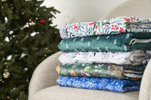 Stacks of wintery fabrics rest on a grey chair, with a christmas tree in hte background.