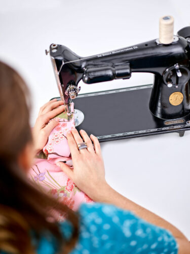 A women with brown hair is sewing pink fabric through a black sewing machine on a white surface.