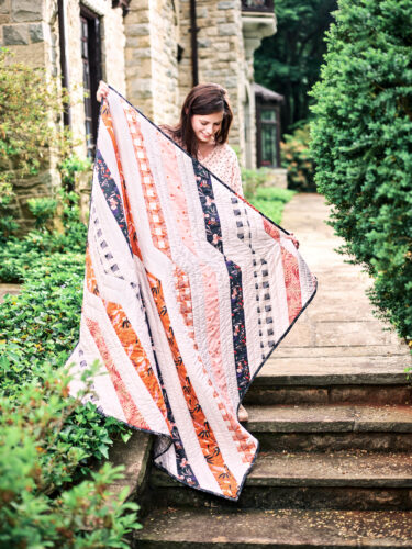 A woman holds a white, blue, and orange quilt while standing on outdoor steps.