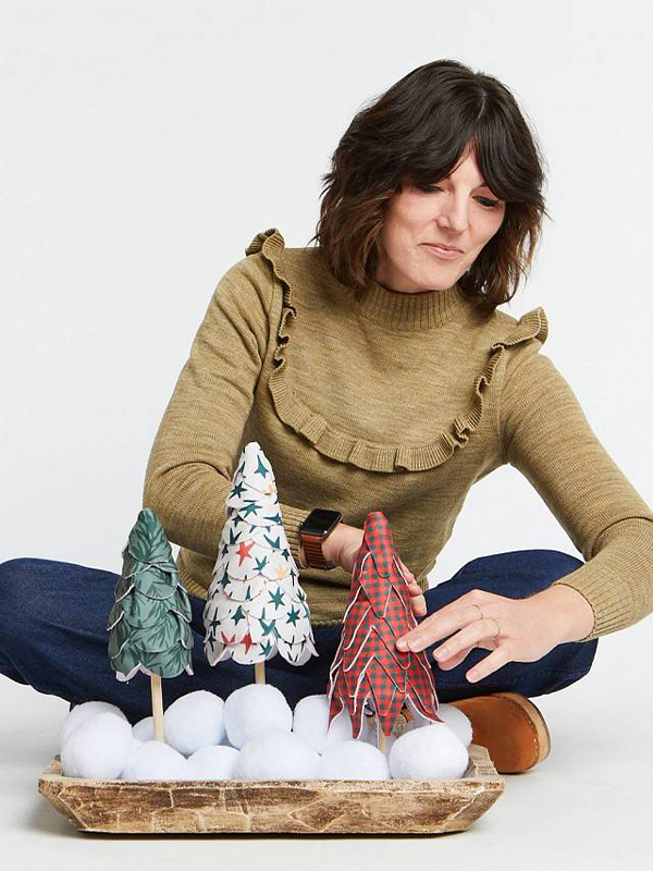 A woman wearing dark blue jeans and a dark yellow long-sleeved top sits on a white floor styling a tray with three trees made by layering small pieces of fabric. One tree has a green design with dark green fir boughs, one tree has a white design with red, white, blue and yellow stars and one tree has a tiny red and dark green gingham design.