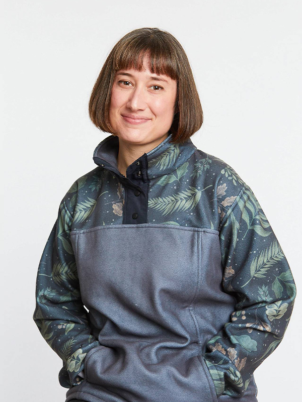 A woman wears a pullover that is textured gray. The pullover’s yoke and sleeves are a slightly darker gray and small cream and orange flowers and green leaves float through the design. She is smiling at the camera, sitting down and in front of a white background.