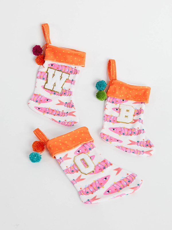 Three small stockings lay on a white surface. The stocking cuffs are orange with small yellow dots and the rest of the stockings feature a design with pink sardines wrapped in colorful small holiday lights on a white background. Each stocking has two jewel-toned yarn pom poms at the top left.