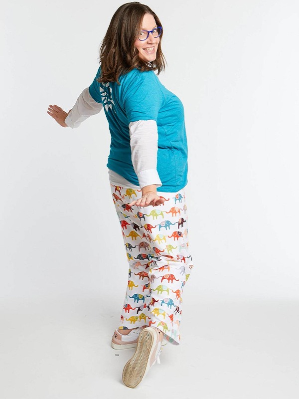 A woman is standing on a white floor and against a white background and is turning around to look at the camera. She is smiling and wearing a turquoise shirt and lounge pants featuring a design with jewel-toned elephants in blue, yellow, orange, red and more, on a white background.