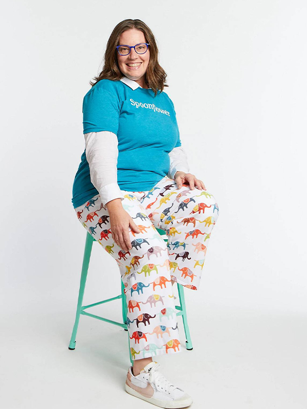 A woman sits on a mint-colored chair against a white floor and background. She is smiling at the camera and wearing a turquoise shirt and lounge pants featuring a design with jewel-toned elephants in blue, yellow, orange, red and more, on a white background.