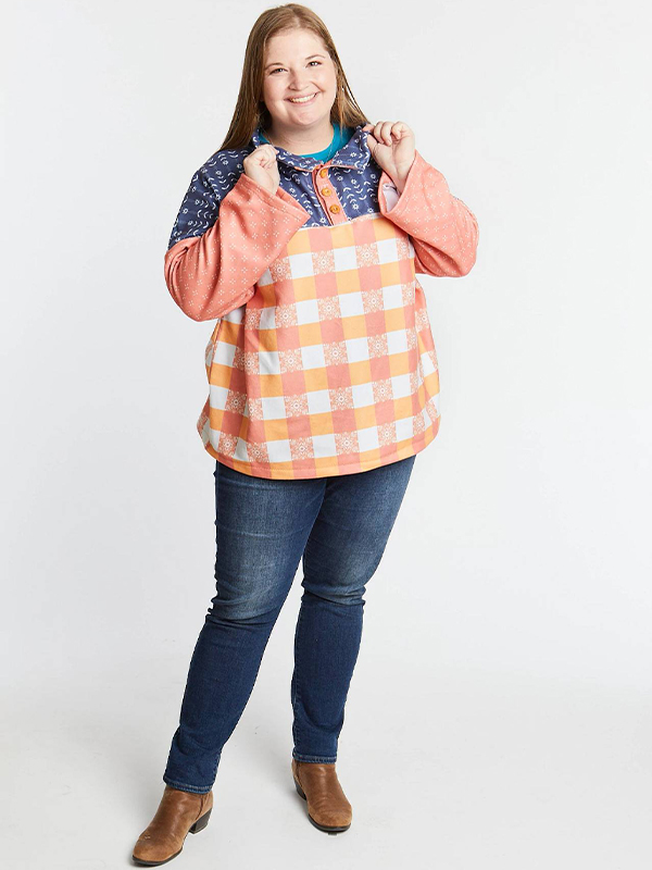 A woman wears a pullover and jeans, each hand is holding onto one side of the pullover’s collar. The fabric designs on the pullover are a peach-and-white gingham, a peach with small white x’s and a navy design with small white flowers. She is smiling at the camera and standing on a white surface and is in front of a white background.