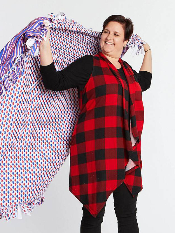 A woman stands in front of a white background and is about to wrap a blanket around her shoulders. She is wearing a long black-and-red vest, black shirt and and black leggings. The designs on the blanket are purple, orange and white.