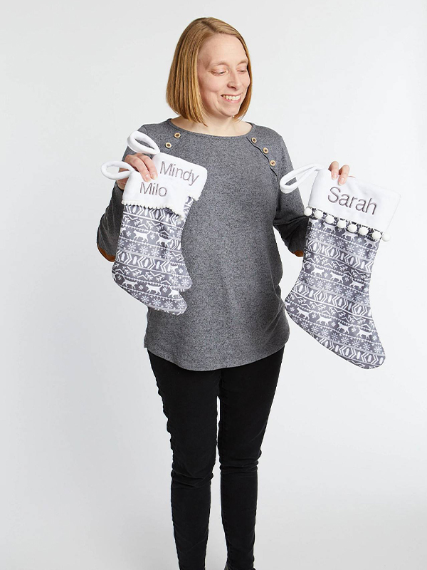 A woman wearing a gray sweater and jeans holds up two small stockings, one in each hand. One stocking says Milo in gray at the top and the other says Mindy, also in gray. The bottom part of the stocking features a repeating Fair Isle design with small white cats, white snowflakes and white trees on a gray background.
