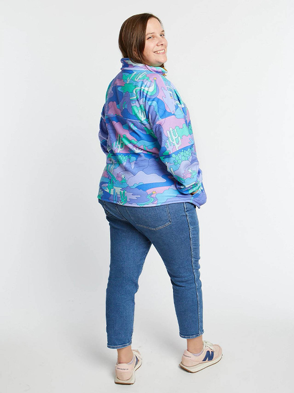 A woman is wearing a jeans and a pullover featuring a turquoise, light blue and purple Arizona landscape design. She is turned around and smiling at the camera and standing on a white surface and against a white background.