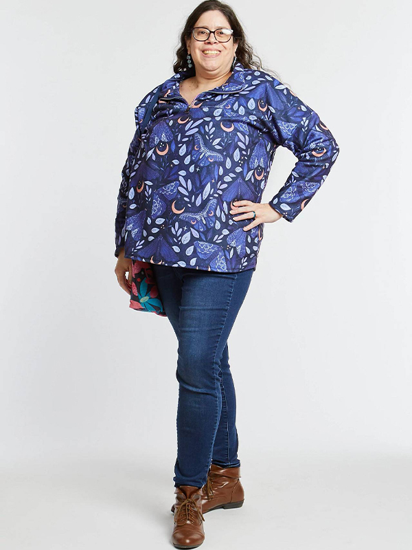 A woman is smiling at the camera and wearing a dark blue pullover with dark purple and lavender moths flying around a small sliver of a golden half moon. She is wearing jeans and standing on a white surface and in front of a white background. Her left hip is on her hip.