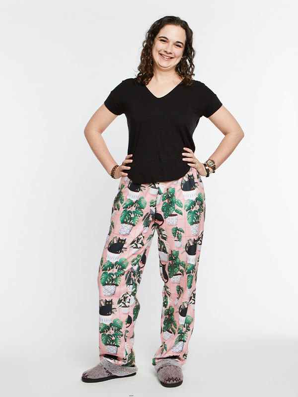 A woman stands in front a white background wearing pink loungepants with a repeating design featuring black cats and green plants in a white planter. Her hands are on her hips and she is smiling at the camera.